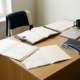 Moderner Schreibtisch mit Lederkladde, Rechnungen, Produktkatalogen und Laptop im warmen Bürolicht.