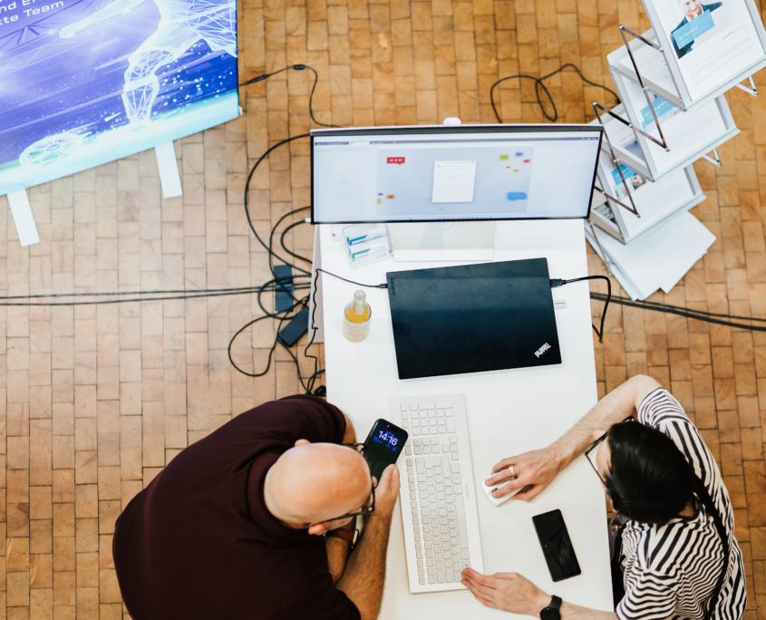 Draufsicht auf unseren Messestand beim Customer Centricity Forum 2026: Kay Frosinini, Senior Consultant & Leitung Marketing und Tobias Dinter, Teamleiter Entwicklung & Support, sitzen sich an der Theke gegenüber und agieren am Monitor.