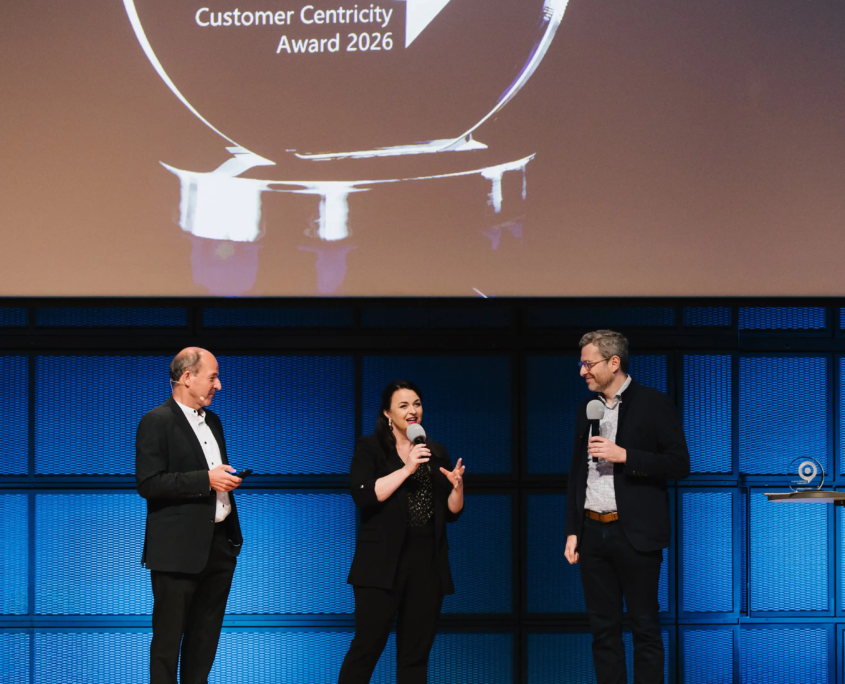 Aufnahme einer weiblichen und zwei männlicher Speaker des Customer Centricity Forums 2026 vor der blau beleuchteten digitalen Wand des Zentrum für Kunst und Medien in Karlsruhe. Über den Personen eine Projektorleinwand mit der Abbildung des Customer Centricity Award 2026.