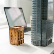 Handgefertigtes Holzmodell eines Bürogebäudes neben einem gläsernen Wolkenkratzer-Miniatur auf weißem Schreibtisch.