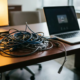 Verwirrtes Knäuel aus Ethernet-Kabeln und USB-Steckern auf einem Büroschreibtisch neben einem Laptop, ein Kabelende hängt lose herab.