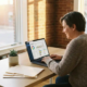 Selbstständige Unternehmerin am Holzschreibtisch mit CRM-Dashboard auf Laptop, warmes Abendlicht, Zimmerpflanze und Ordner im Hintergrund.