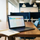 Laptop auf Konferenztisch zeigt farbiges Kontakt-Pipeline-Dashboard, daneben Notizbuch und Stift, modernes Büro im Hintergrund.