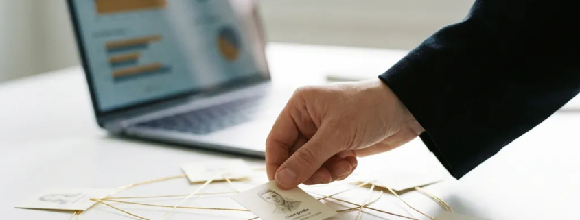Hand fügt letztes Kundenprofil-Karte in kreisförmiges Netzwerk ein, verbunden durch goldene Fäden, modernes Büro mit Laptop im Hintergrund.