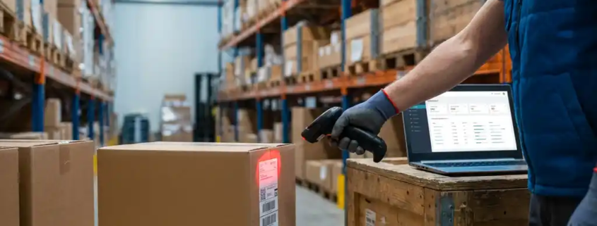 Lagerarbeiter scannt Barcode auf Karton mit Handscanner, im Hintergrund geordnete Metallregale mit Inventar, Laptop mit Dashboard im Vordergrund.