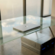 Laptop auf Glasschreibtisch neben physischem Server, Bürofenster spiegelt Wolken im Bildschirm, silber-blaue Farbtöne.