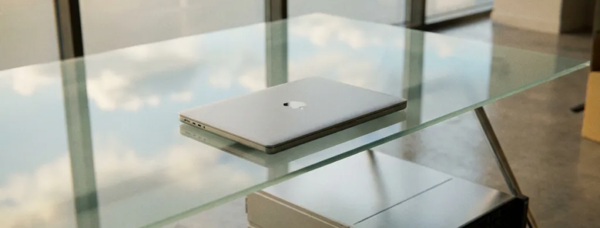 Laptop auf Glasschreibtisch neben physischem Server, Bürofenster spiegelt Wolken im Bildschirm, silber-blaue Farbtöne.
