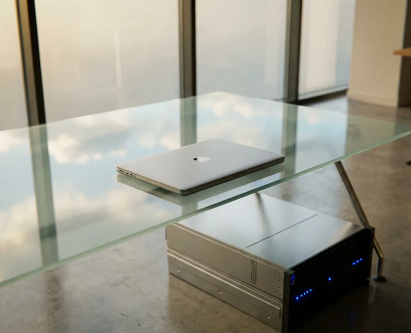 Laptop auf Glasschreibtisch neben physischem Server, Bürofenster spiegelt Wolken im Bildschirm, silber-blaue Farbtöne.