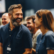 Bild von Veranstaltungsbesuchern beim entspannten Networking. Im Vordergrund ein lachender Mann in dunkelblauem Hemd mit kurzen Haaren und grau meliertem Vollbart und eine ebenfalls lachende Frau in blauer Bluse und braunen glatten langen Haaren, die sich ansehen. Links und rechts über dem Bild liegt ein transparentes technisches Muster in bunten Farben.