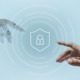 Bild einer Roboterhand und einer Menschenhand, die von links und rechts in das Bild hereinragen und mit dem Zeigefinger auf einen weißen Icon in Form eines Schutzschildes mit einem Schloss in der Mitte zeigen. Von dem Icon gehen dezente weiße kreisförmige Intervalle aus.