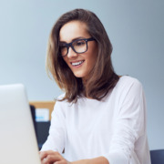 Bild einer jungen braunhaarigen Frau mit scharzer Brille und weißem Shirt, die enspannt lächelnd auf ihrem weißen Laptop tippt. Im Hintergrund ist etwas unscharf Büroeinrichtung zu erkennen.