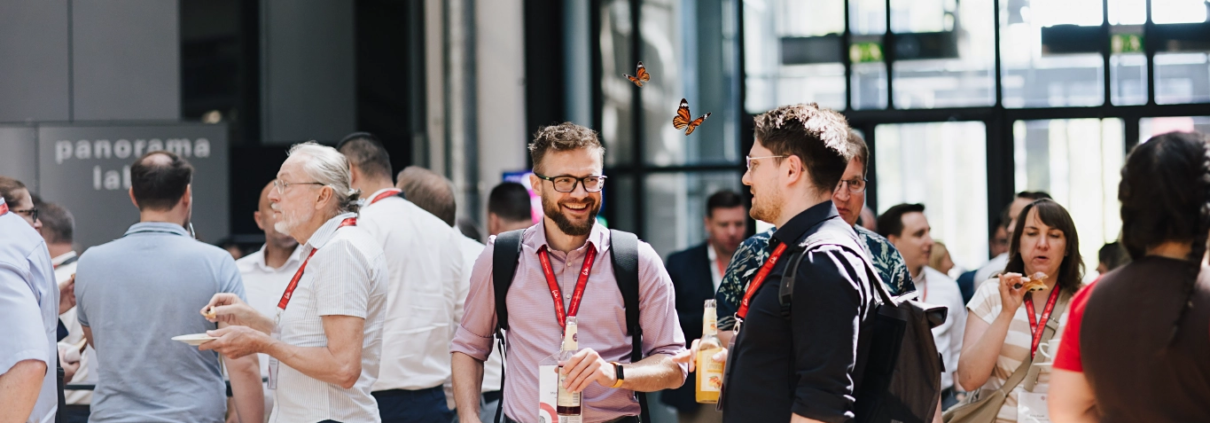 Aufnahme von Besuchern des Customer Centricity Forums 2026 beim Smalltalk und Snacken im Foyer des ZKM, Zentrum für Kunst und Medien in Karlsruhe. Zwischen den Besuchern flattern zwei kleine rot-schwarze Schmetterlinge herum.
