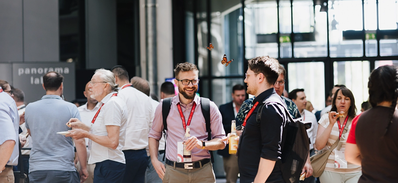 Aufnahme von Besuchern des Customer Centricity Forums 2026 beim Smalltalk und Snacken im Foyer des ZKM, Zentrum für Kunst und Medien in Karlsruhe. Zwischen den Besuchern flattern zwei kleine rot-schwarze Schmetterlinge herum.