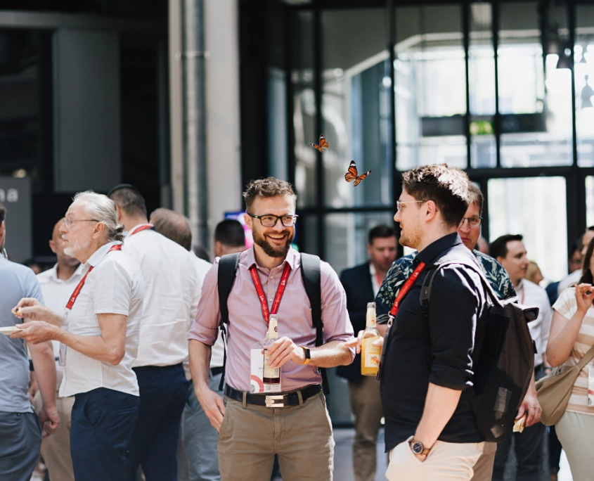 Aufnahme von Besuchern des Customer Centricity Forums 2026 beim Smalltalk und Snacken im Foyer des ZKM, Zentrum für Kunst und Medien in Karlsruhe. Zwischen den Besuchern flattern zwei kleine rot-schwarze Schmetterlinge herum.
